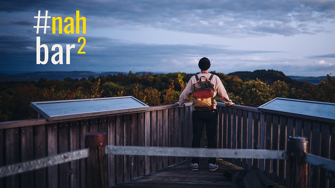Eine Person mit einem Rucksack steht auf einer hölzernen Aussichtsplattform und blickt in der Abenddämmerung auf eine bewaldete Landschaft. Der Text "#nahbar²" ist in Gelb und Weiß auf der linken Seite des Bildes zu sehen.
