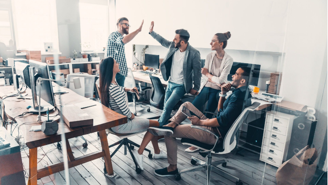 Eine Gruppe von fünf jungen Erwachsenen in einem modernen Büro, die sich locker unterhalten und High Fives geben. Einige sitzen auf Stühlen, andere stehen. Der Arbeitsbereich ist mit Schreibtischen, Computern und Büromaterial ausgestattet.