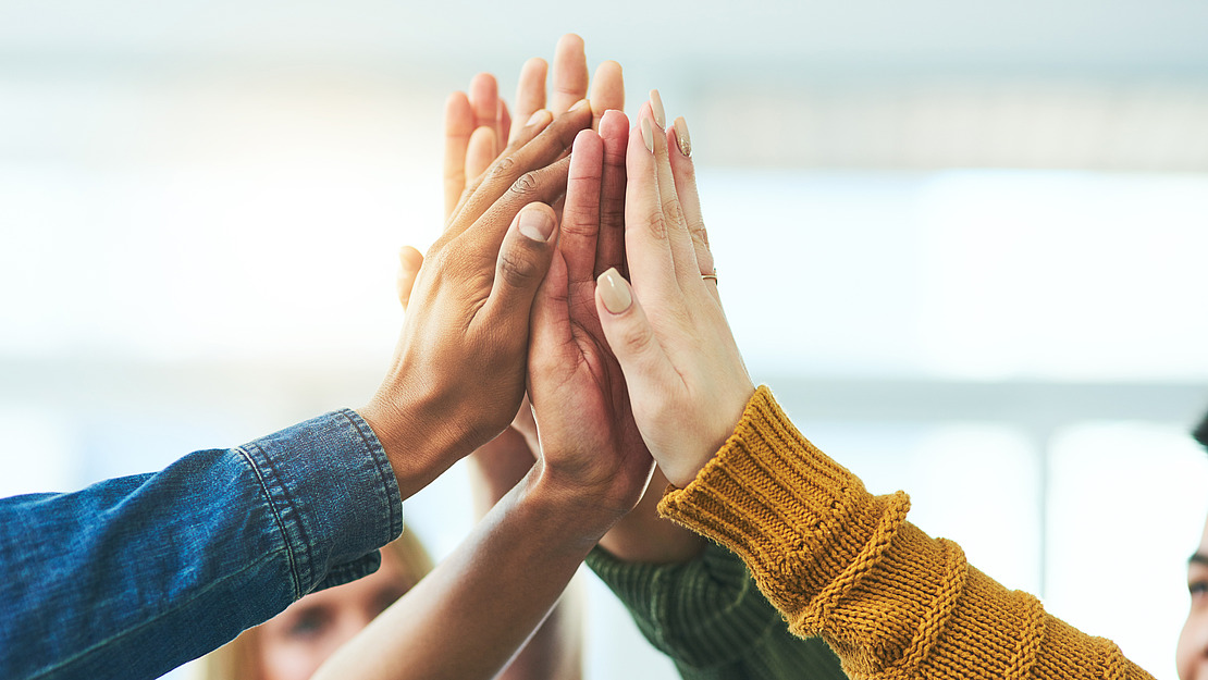 Eine Gruppe unterschiedlicher Hände, die sich zu einem High-Five zusammentun, symbolisiert Teamwork und Einheit. Jede Person trägt unterschiedliche Kleidung, darunter eine Jeansjacke und einen gelben Pullover. Der Hintergrund ist leicht verschwommen und hell.