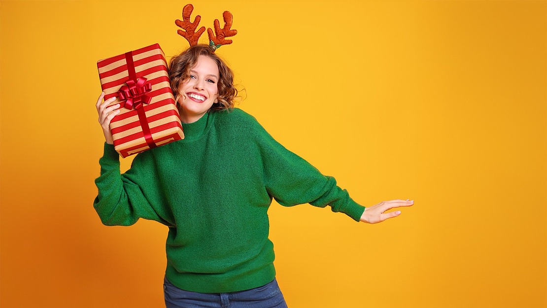 Eine lächelnde Person mit Rentiergeweih und grünem Pullover hält ein eingepacktes Geschenk mit einer roten Schleife auf der Schulter und steht vor einem hellgelben Hintergrund.