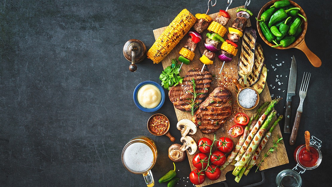Eine Draufsicht auf ein Holzbrett mit gegrillten Steaks, Gemüse, Mais, Pilzen, Tomaten und Dip-Saucen, umgeben von einem Glas Bier, Paprika und Gewürzen auf dunklem Hintergrund.
