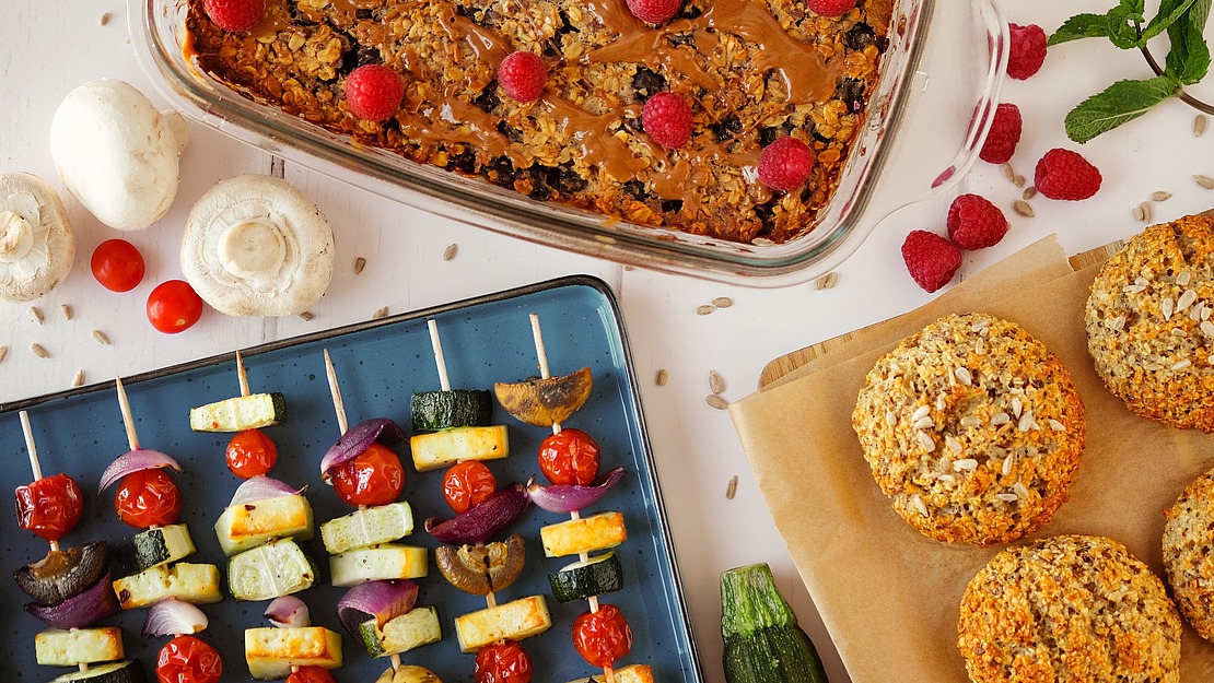 Ein gebackenes Haferflocken-Gericht mit Himbeeren, Gemüsespieße mit Tomaten, Zucchini, Zwiebeln und Pilzen sowie vier runde, herzhafte Bratlinge auf Backpapier, umgeben von frischem Obst und Gemüse.