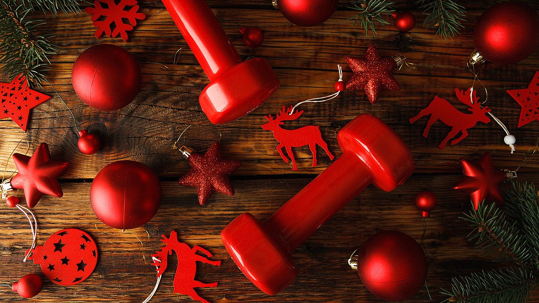 Rote Hanteln sind von festlicher Weihnachtsdekoration umgeben, darunter rote Ornamente, Sterne und Rentier-Ausschnitte auf einem Holzhintergrund mit immergrünen Zweigen.