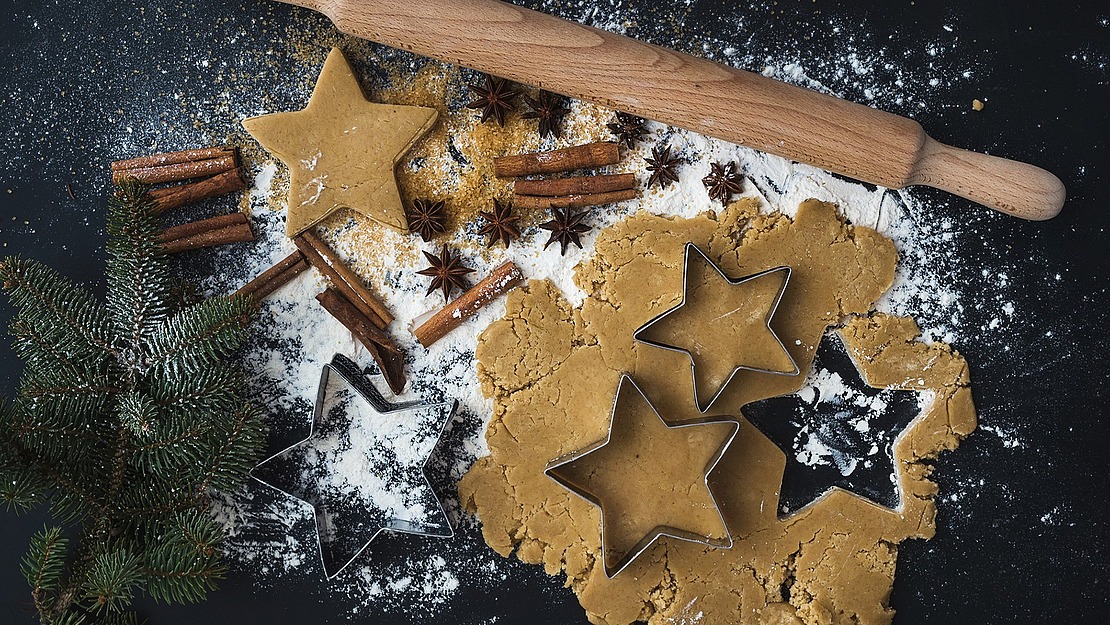 Auf einer bemehlten Fläche ausgerollter Plätzchenteig mit sternförmigen Ausstechern, einem Nudelholz, Zimtstangen, Anissternen, Tannenzweigen und gestreutem Mehl, die an eine festliche Weihnachtsbäckerei erinnern.