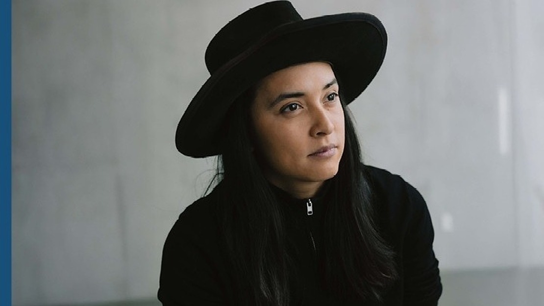 Eine Person mit langen dunklen Haaren, die einen breitkrempigen schwarzen Hut und ein schwarzes Oberteil trägt, schaut vor einem schlichten, hellgrauen Hintergrund nachdenklich in die Ferne.