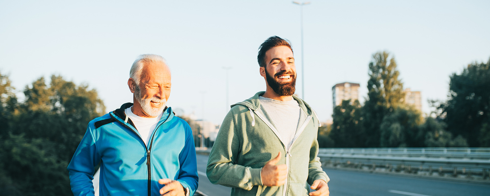 Zwei Männer joggen zusammen über eine Brücke. Einer ist älter, hat weißes Haar und eine blaue Jacke, der andere ist jünger, hat einen Bart und eine grüne Jacke. Sie lächeln und genießen den sonnigen Tag mit Bäumen und Gebäuden im Hintergrund.