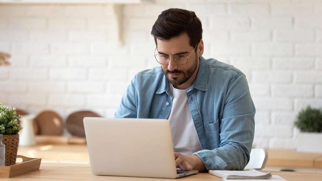Ein Mann mit Brille und Bart sitzt an einem Tisch und arbeitet an einem Laptop. Er trägt ein hellblaues Hemd über einem weißen T-Shirt. Im Hintergrund sind eine weiße Backsteinmauer und zwei kleine Topfpflanzen zu sehen.