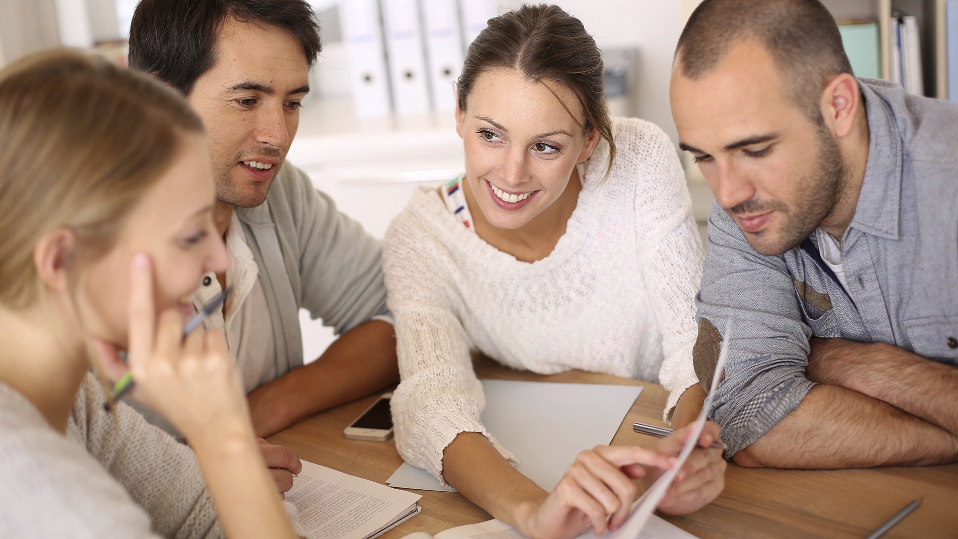 Vier Erwachsene sitzen an einem Tisch, sehen sich Dokumente an und diskutieren gemeinsam. Eine Frau zeigt auf ein Papier, während die anderen zuhören und sich einbringen, was auf eine gemeinsame Sitzung oder ein gemeinsames Studium hindeutet.