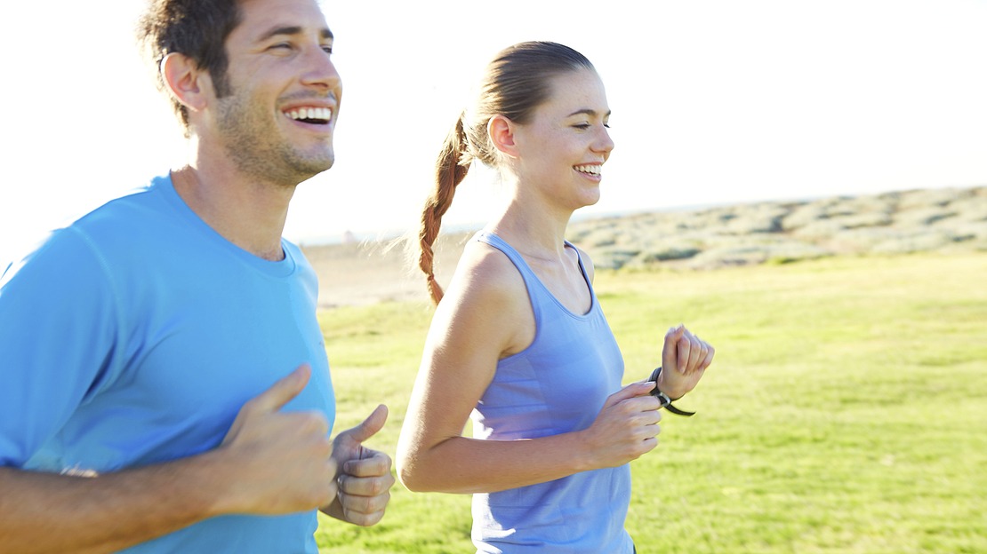 Ein Mann und eine Frau laufen an einem sonnigen Tag im Freien, lächelnd und in hellblauer Sportkleidung. Im Hintergrund sind grünes Gras und eine offene Landschaft zu sehen.
