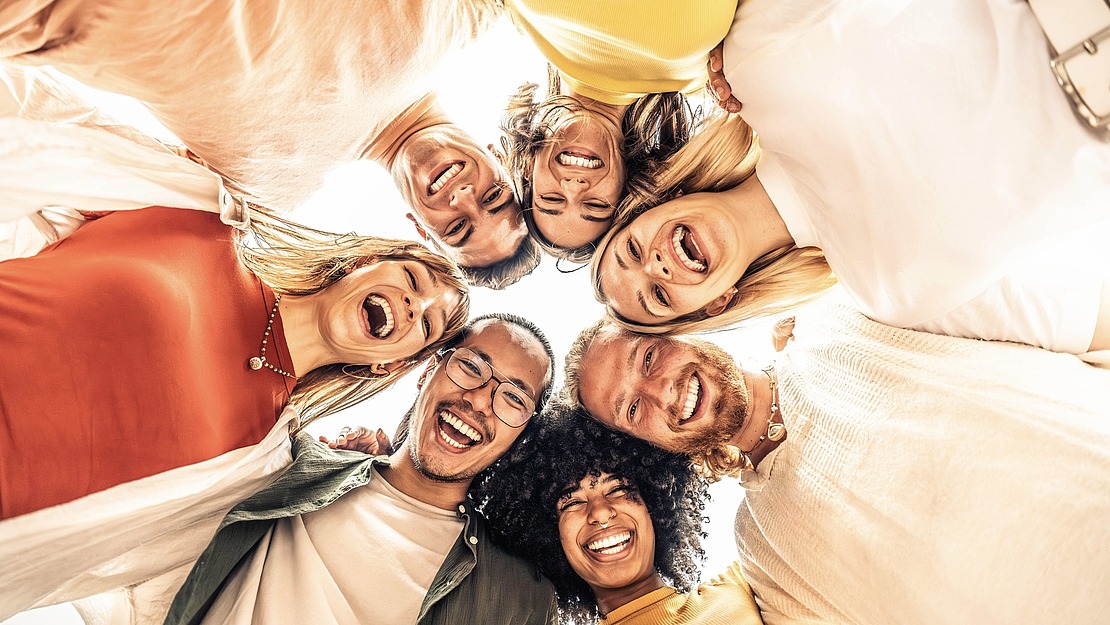 Eine Gruppe von sieben Personen, die in einem Kreis stehen, in die Kamera schauen und glücklich lächeln, mit hellem, natürlichem Licht im Hintergrund.