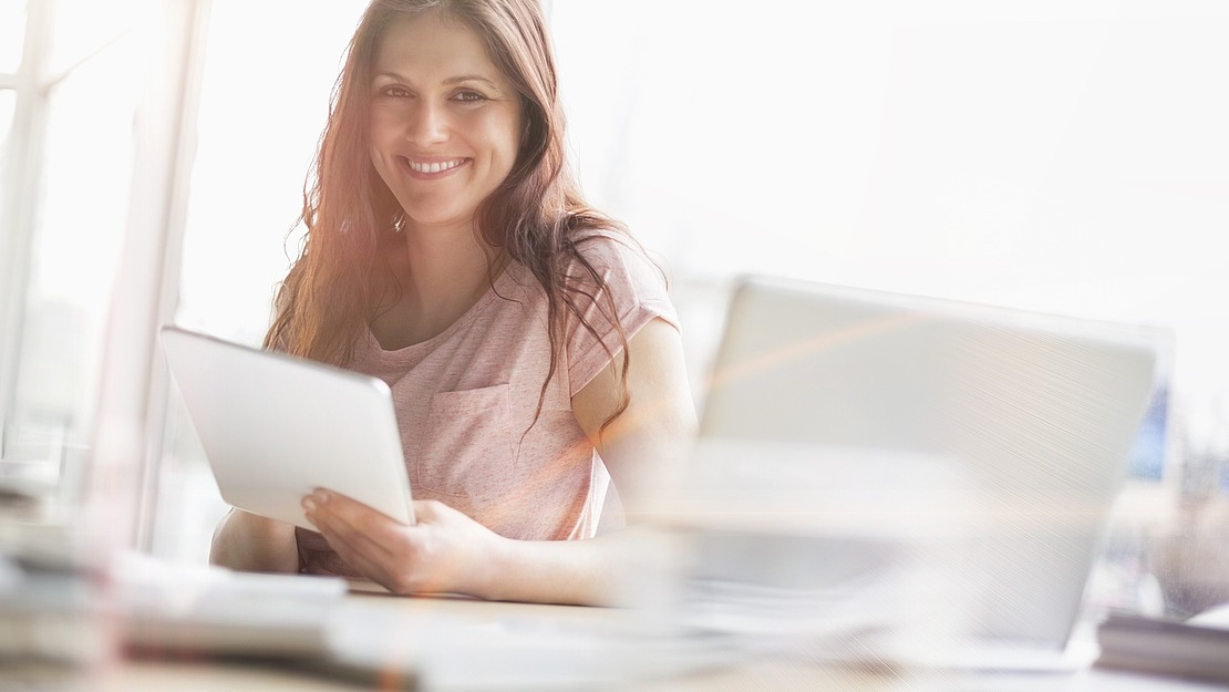 Eine Frau mit langen braunen Haaren, die ein rosa Hemd trägt, lächelt, während sie ein Tablet an einem Schreibtisch hält. Auf dem Schreibtisch befinden sich ein Laptop und Papiere, und die Szene ist durch das natürliche Licht großer Fenster hell erleuchtet.