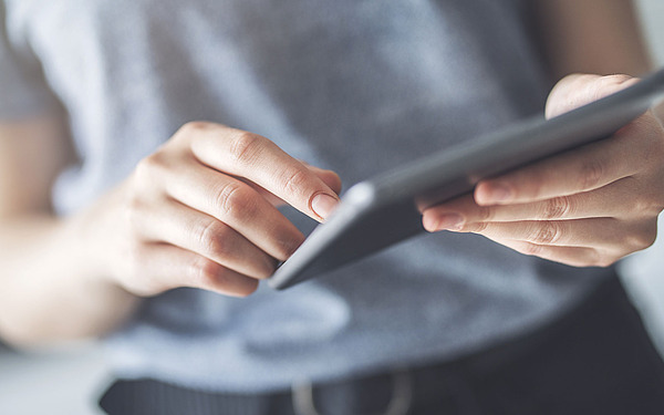 Eine Person in einem grauen Hemd bedient ein Touchscreen-Tablet. Der Fokus liegt auf ihren Händen und dem Gerät, der Hintergrund ist unscharf.