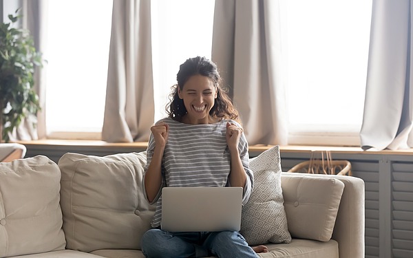 Eine Frau sitzt auf einem Sofa und blickt freudestrahlend auf ihrem Laptop.