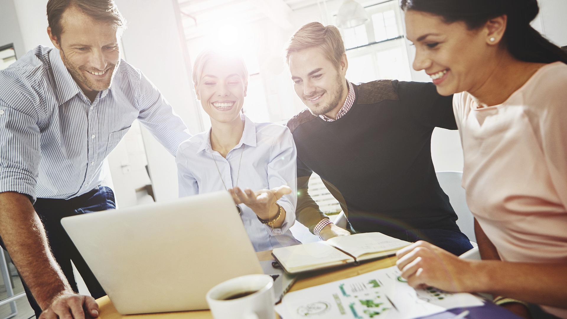 Eine gemischte Gruppe von vier Personen lächelt und schaut in einem hell erleuchteten Büro auf einen Laptop-Bildschirm. Papiere und eine Kaffeetasse liegen auf dem Tisch. Sie wirken kooperativ und engagiert.