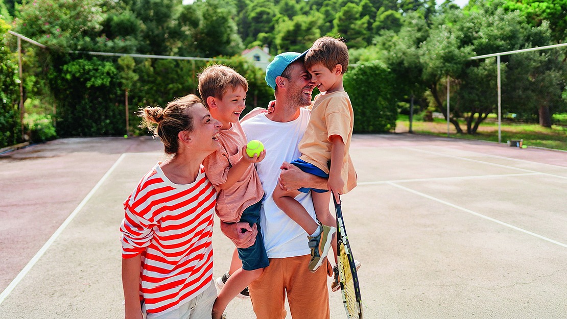 Eine fröhliche Familie auf einem Tennisplatz. Ein Erwachsener in einem gestreiften Hemd lächelt und hält ein Kind im Arm. Ein anderer Erwachsener hält einen Schläger und ein Kind im Arm. Im Hintergrund umgeben Bäume den Platz.