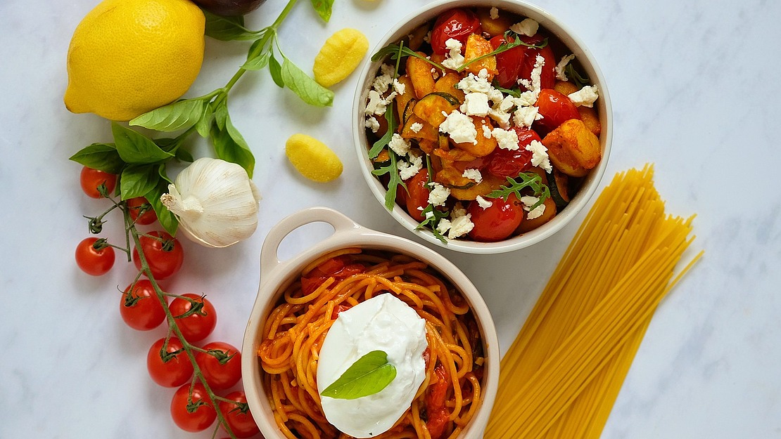 Eine Schüssel Spaghetti mit einem Klecks Käse und eine Schüssel Gnocchi mit Tomaten und Feta, umgeben von frischem Gemüse, rohen Nudeln und Basilikum.