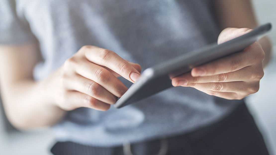 Eine Person in einem grauen Hemd bedient ein Touchscreen-Tablet. Der Fokus liegt auf ihren Händen und dem Gerät, der Hintergrund ist unscharf.